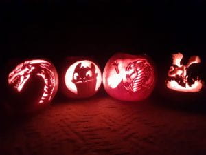 Four carved pumpkins illuminated by a candle. From left to right they are a wingless dragon and its neck, Toothless sticking his tongue out, an epic fire breathing dragon with wings, and the dragon form of Maleficent