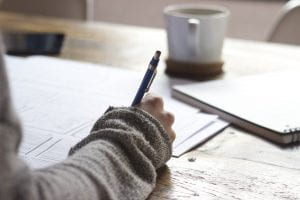 An array of papers are scattered on a table with a sweatered hand taking detailed notes on them. A coffee mug sits nearby